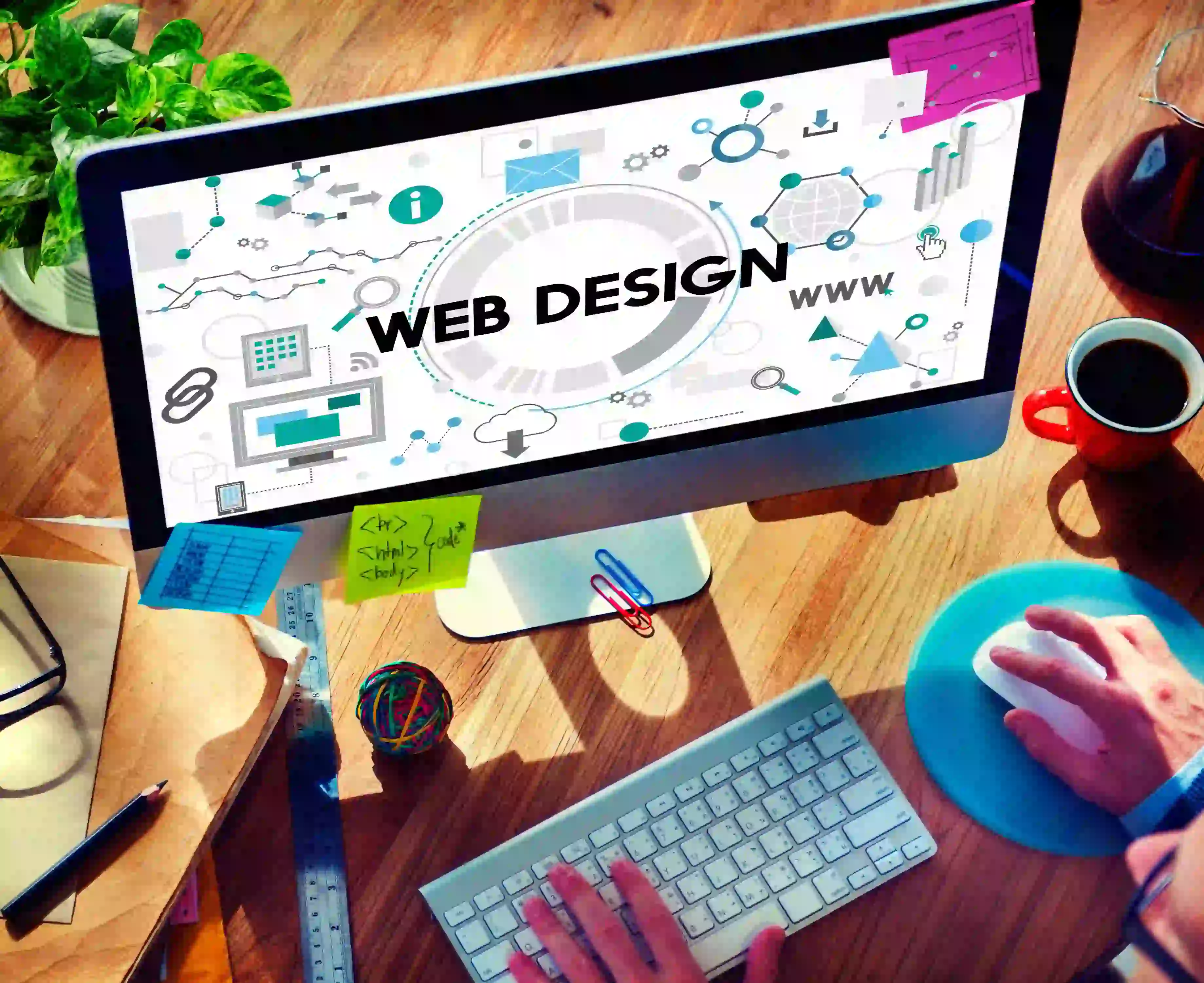 A person works on web design at a computer desk, featuring design tools on the monitor, a keyboard, coffee, notebooks, and scattered stationery.